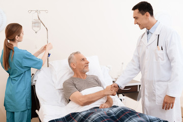 The old man lies on a cot in the medical ward, and next to him stands a doctor. The doctor shakes the old man's hand.
