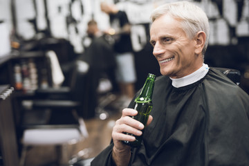 The old man drinks alcohol in the barber's chair in barbershop.