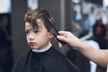 The hairdresser makes a fashionable pretty hairstyle for the boy in a modern barbershop.