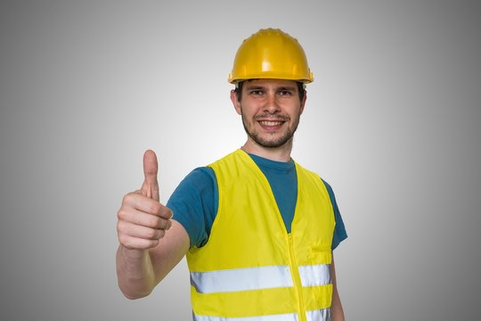 Young happy construction worker in yellow helmet is showing thumbs up gesture.