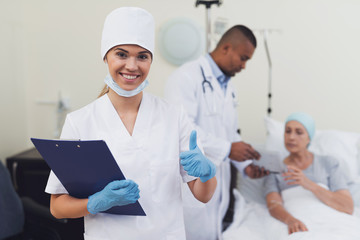 A nurse is posing against the patient's background. She holds a paper tablet in her hands.