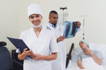 A nurse is posing against the patient's background. She holds a paper tablet in her hands.