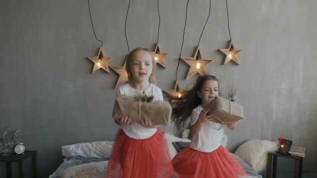 Two little sisters have fun jumping on the bed and holding Christmas gifts in their hands. On a gray wall hangs 6 decorative wooden stars that shine with lights