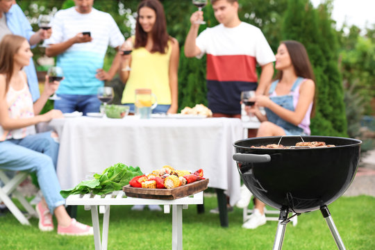 Barbecue grill, table with prepared vegetables and blurred people on background
