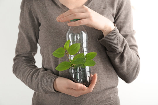 Young woman with plastic bottle used as container for growing plant against light background