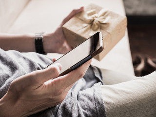 Close up of casual man's hands using smart phone and holding gift box.