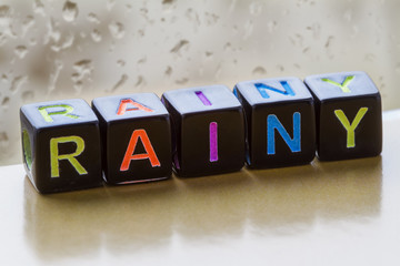 Rainy weather. The inscription rainy from toy cubes with colored letters on the background of a window with raindrops. Closeup, selective focus