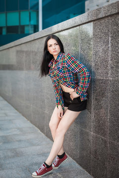 Young hipster woman with long hair leaning against the wall in the street, wearing colorful shirt in cage and black shorts on the urban background