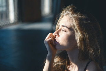 Sideview close up shot of pretty female with loose voluminous hair and clean skin sitting in bedroom in the morning, having pensive or dreamy look, making plans for day, sun shining on her face