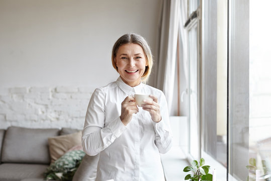 Positive cheerful 60 year old woman medical expert with gathered hair standing by large window in modern apartment interior, having coffee break while working from home, consulting patients