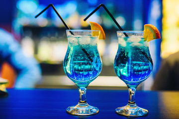 two glasses of blue cocktail Martini with orange and ice cubes and straws on bar