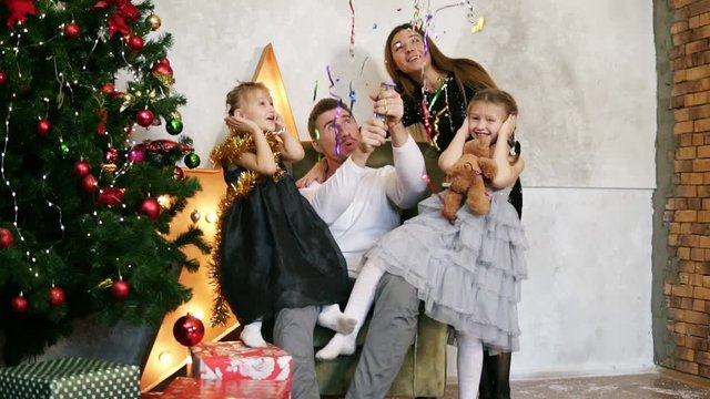 Family of four having fun with petards sitting by the decorated Christmas tree. Happy father, mother and two daughters celebrating Christmas together