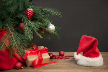 Christmas holiday background. Gifts with a red ribbon, Santa's hat and decor under a Christmas tree on a wooden board. Close up. Copy space on chalkboard.