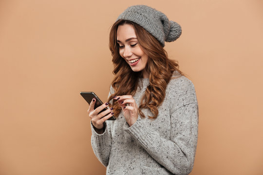 Young smiling brunette woman in woolen hat and sweater typing message on mobile phone