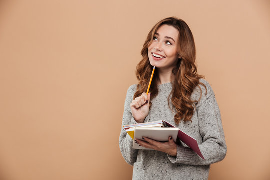 Portrait of happy thinking female student in warm sweater holding pencil and notebooks, looking aside