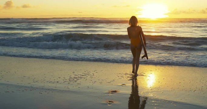 Beautiful Young Woman in a Swimsuit Walks into the Ocean while Carrying Surfboard. She is Looks Gorgeous and Slim, Has Blonde Hair. Big Oceanic Waves and Sunset in the Background.