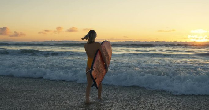 Beautiful Young Woman in the Swimsuit Walks into the Ocean while Carrying Surfboard. She is Looks Gorgeous and Slim, Has Blonde Hair. Big Oceanic Waves and Sunset in the Background.