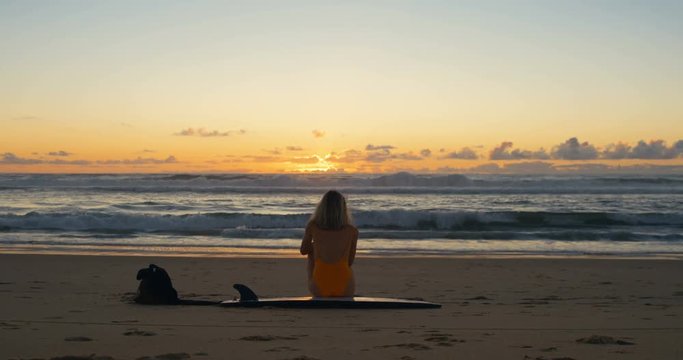 Beautiful Young Woman in Swimsuit Sitting on the Beach on Her Surfboard Looking at the Big Oceanic Waves and Watching the Sunset.