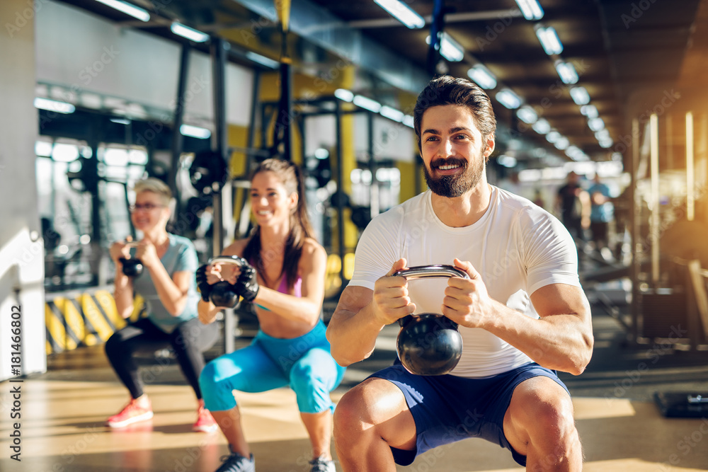 Canvas Prints Attractive handsome bearded man holding kettlebell and doing squats in a fitness group with two girls in the modern gym.