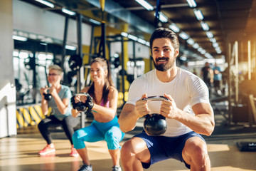 Fototapeta na wymiar Attractive handsome bearded man holding kettlebell and doing squats in a fitness group with two girls in the modern gym.