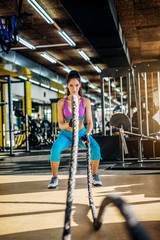 Attractive strong fitness woman doing battle rope exercise in the modern sunny gym.