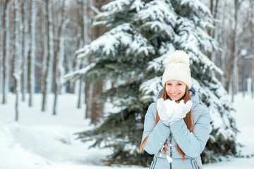 A Girl Wearing Warm Winter Clothes And Hat Blowing Snow In Winter Forest, horizontal. Model with a beautiful smile near the Christmas tree.