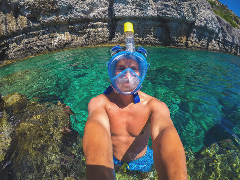 Selfie photo of a young healthy muscular man with a snorkelling mask standing in the turquoise exotic sea near the rocks for summer holidays.