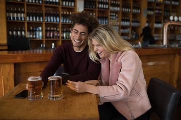 Couple using mobile phone while having glass of beer in bar