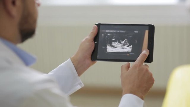Doctor in white medical robe shows pictures of ultrasound to pregnant woman on a tablet