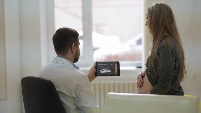 A doctor in a white medical dressing gown shows a pregnant woman on a tablet the pictures of an ultrasound with the problem of a baby fetus