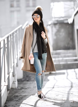 All fashion outfit details. Fashionable woman wearing coat, ripped jeans with sneakers.