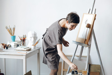 Female artist in her spacious white studio working with watercolor painting dipping a brush into...