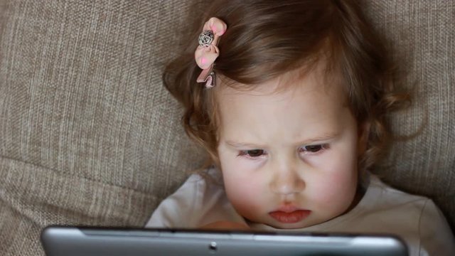 Funny child looks at the screen and plays downloaded application on tablet laptop, computer A little cute girl lies in sofa in a living room, looking cartoon and playing the game. close-up