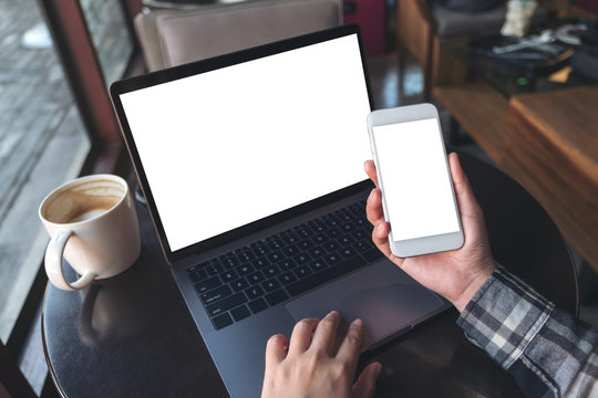 Mockup top view image of business woman holding white mobile phone with blank screen with laptop and coffee cup on wooden table in cafe