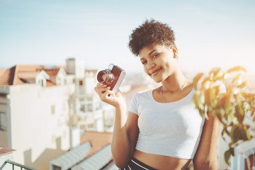 Dazzling young smiling Brazilian female with retro film camera on the roof of Lisbon residential house; cheerful black curly girl with vintage photo camera on the balcony with cityscape in background