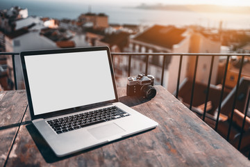 Mock-up of empty screen of modern notebook standing on wooden table outdoor on balcony; laptop with blank white screen template and vintage film photo camera near, defocused cityscape in background - Powered by Adobe