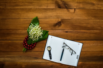 Notepad, clock, glasses, pen and lily of the valley with beads lie on a wooden background, place for your text. View from above.