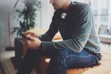 Young hipster wear casual clothes and headphones typing fingers on smartphone screen at modern loft studio.Horizontal.Blurred background.