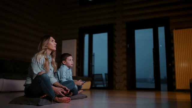 Beautiful mother with her son playing video games on gaming console in the living room of his house. Happy family in a country house.