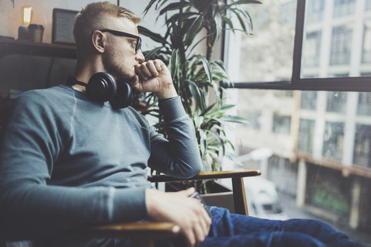 Pensive young man wearing glasses casual clothes.Man sitting in vintage armchair modern loft studio and relaxing whith headphone music.Panoramic windows on blurred background.Horizontal.