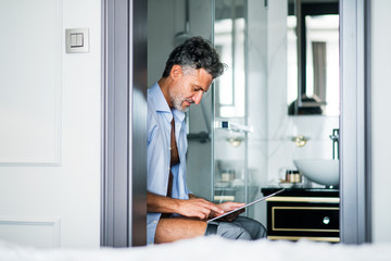 Naklejka na ściany i meble Mature businessman with tablet in a hotel room bathroom.