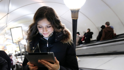 Young attractive Caucasian girl looking at the screen of the tablet and holds it with his fingers, rises on the escalator out of the subway