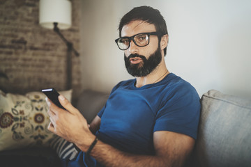 Young bearded man wear casual clothes and eye glasses typing fingers on smartphone screen at modern loft home.Horizontal.Blurred background.