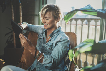 Smiling young woman wearing casual clothes and using electronic device while spending relax time home livingroom.Attractive woman owner communicating on digital tablet.Blurred background.