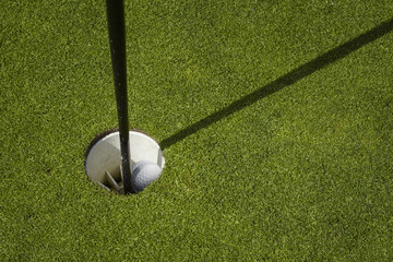 Golf ball sit inside cup on golf course putting green with flag.