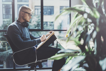 Skilled designer caucasian man drawing abstract sketch with pen.Art work process.Creative hobby.Noting ideas in copy book on workspace desk with papers in modern studio with panoramic windows.Blurred.