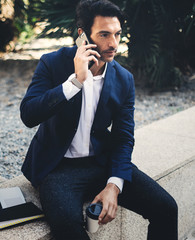 Pensive elegant businessman using contemporary smartphone while taking rest during work day.Holding hands take away coffee cup.Blurred background. Vertical.