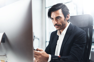 Young elegant man working at sunny office on desktop computer while sitting at wooden table.Blurred background,horizontal.