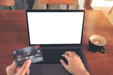 Mockup image of a hand holding credit cards while using and typing on laptop with blank green screen and coffee cup on wooden table in modern loft cafe