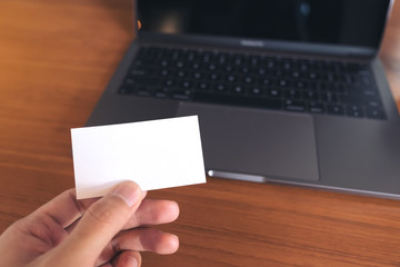 A hand holding white empty business card with laptop and coffee cup on wooden table in office
